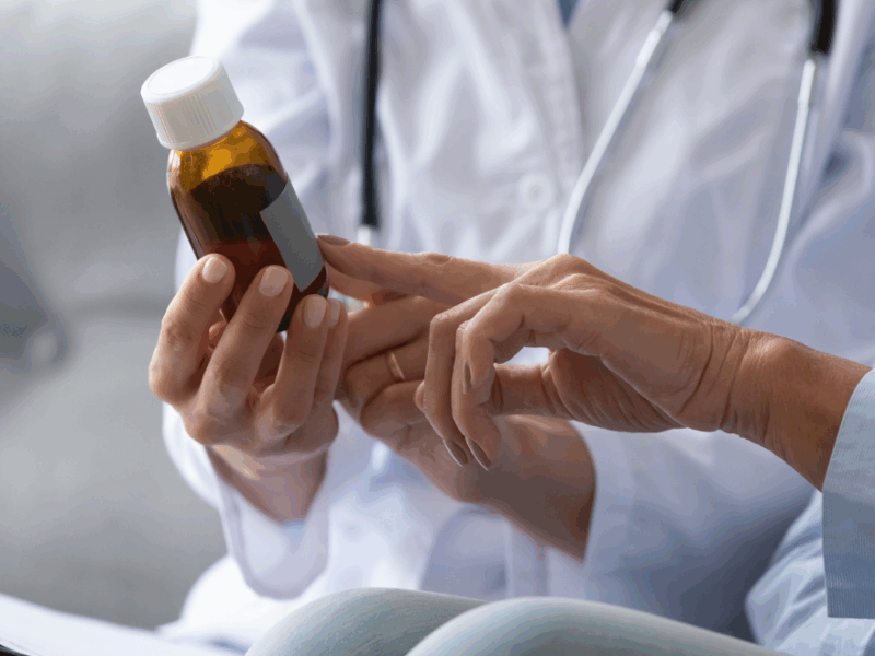 Close up of a female doctor's hands as she holds up a prescription bottle and shows it to a female patient.