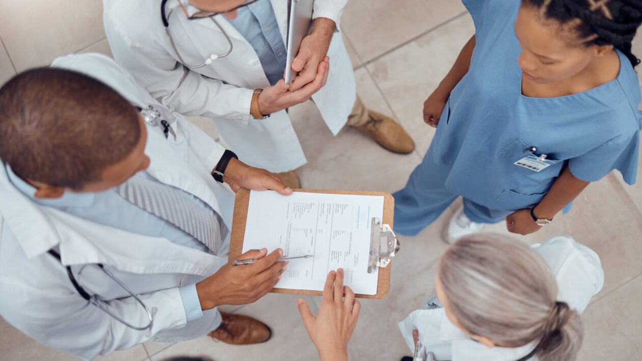 A birds-eye view of a group of NHS professionals in uniforms (doctors, nurses) gathered in a circle,focused on a document one of them is holding.