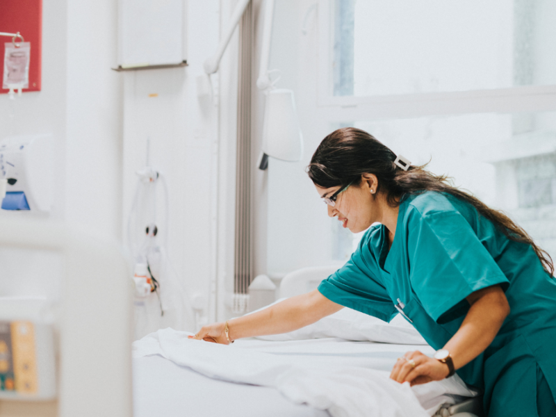 A healthcare worker making a bed in a clinical room, overlaid with the Hub's graphic device - curved colour bands and the title: Case Study – Strengthening linen and laundry services across the East of England through procurements worth over £100 million.