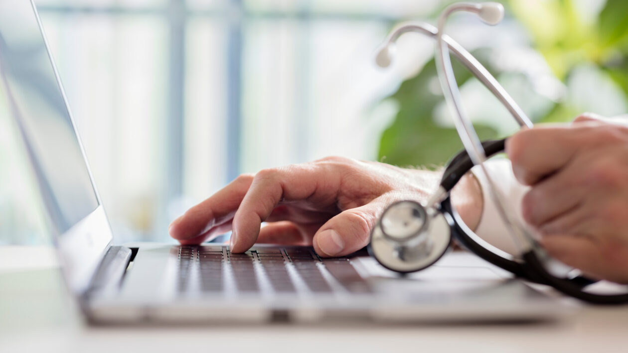 Crop close up of female GP or physician sat at a desk in hospital typing on a laptop.