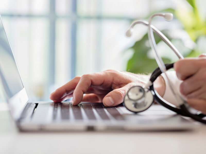 Crop close up of female GP or physician sat at a desk in hospital typing on a laptop.
