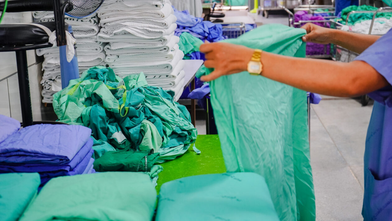 Person folding green surgical linen on a work surface in a hospital laundry or sterilisation area, with stacks of folded white towels and piles of green and blue clinical fabric in the background.