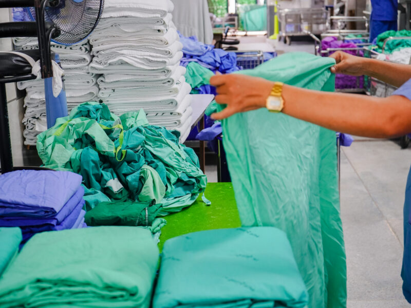 Person folding green surgical linen on a work surface in a hospital laundry or sterilisation area, with stacks of folded white towels and piles of green and blue clinical fabric in the background.