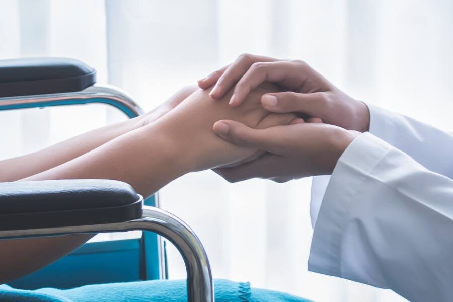 A close up of a doctor in a white coat with a stethoscope around her neck crouching down to a young patient in a wheelchair holding hands.