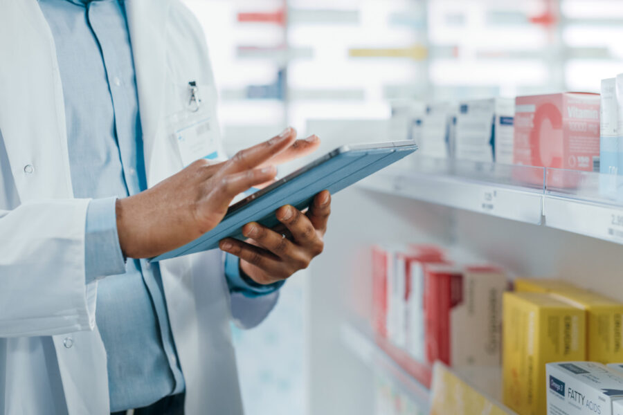 A pharmacist standing in front of shelves of medicine taking inventory with a tablet, with the focus on his hands.