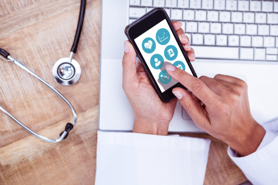 Hands holding a mobile phone with healthcare apps on it. In the background there is a laptop and a stethoscope.