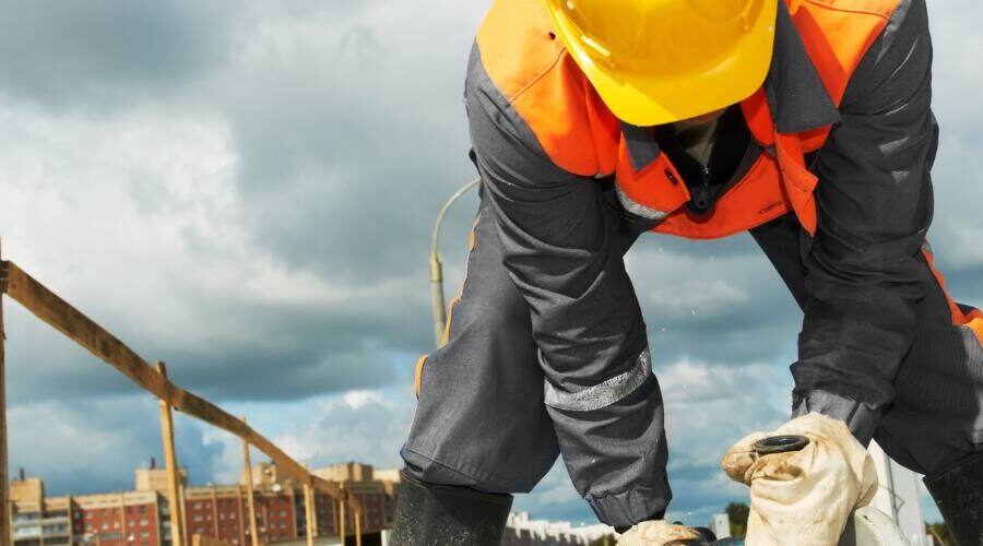 A construction worker is crouching down on top of a building site using a saw.