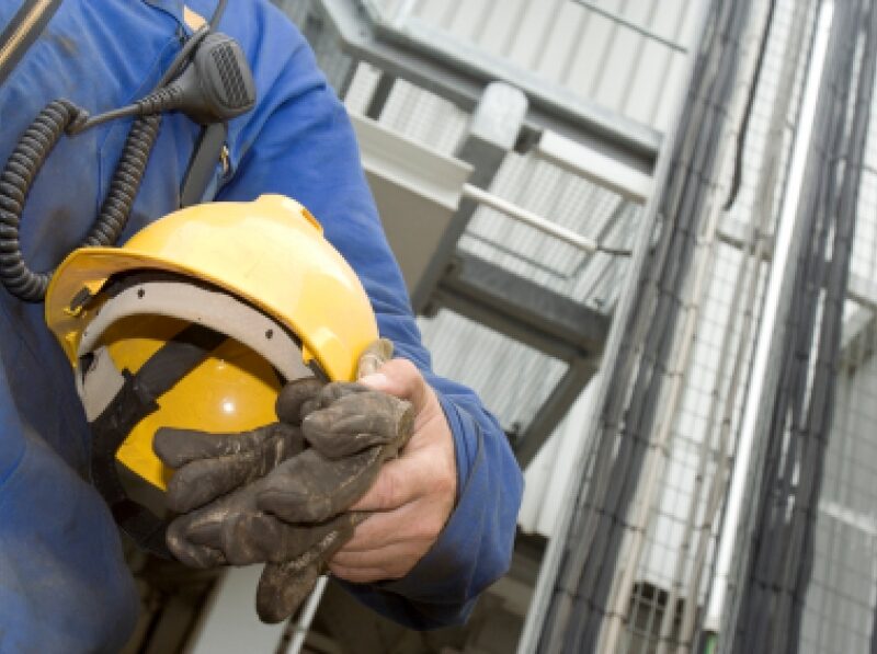 A close up of a person standing in front of a building site and holding a hard hat under their arm.