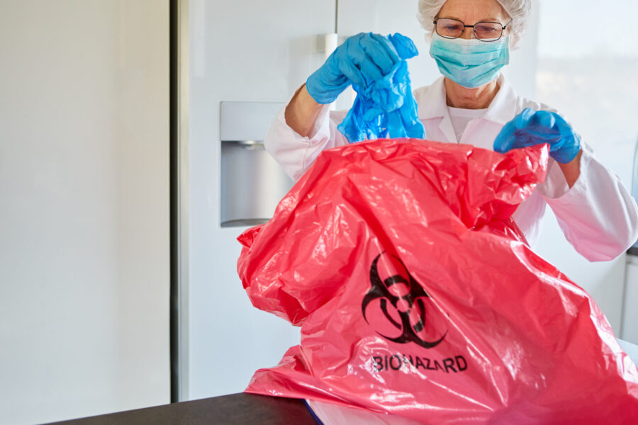 An older female cleaner wearing a facemask and gloves drops used PPE into a biohazard bag